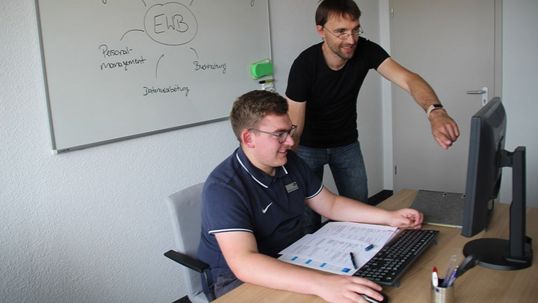 Maximilian Reinisch ist zurzeit Azubi bei EWB und sitzt am Computer, Sebastian Schulze vom Bereich Datenmanagement steht neben im und zeigt auf den Bildschirm